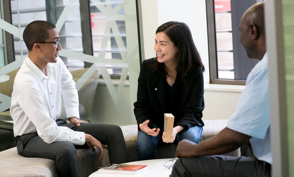 Business students talking Two male and one female business students sitting on a circular bench indoors, talking.